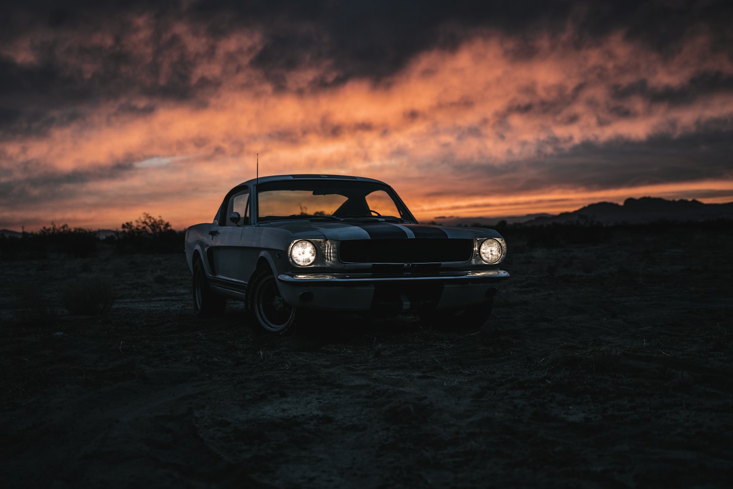 A classic Mustang muscle car parked at twilight with glowing headlights.
